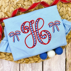 Könnte beinhalten: Ein hellblauer Baby-Strampler mit einem roten und blauen gestickten Buchstaben "H" und zwei roten, weißen und blauen Schleifen. Der Strampler ist mit drei Bommeln verziert, zwei blauen und einem weißen.
