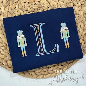May include: A navy blue t-shirt featuring an embroidered letter L in light blue and gold, accompanied by two nutcracker figures. The nutcracker figures are dressed in blue and gold uniforms with red accents.
