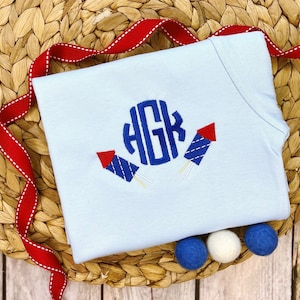 Könnte beinhalten: Ein hellblaues T-Shirt mit einem gestickten Feuerwerksmotiv in Blau und Weiß und einem Monogramm der Buchstaben "HGR" in einem Kreis.