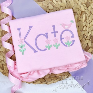 May include: A light pink shirt embroidered with the name "Kate" in lavender letters. The "K" and "E" are decorated with pink tulips and green leaves. A pink bird sits above the "E". The shirt has a ruffled hem.