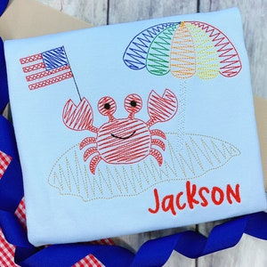 Könnte beinhalten: Ein hellblaues T-Shirt mit einem gestickten roten Krebs, der einen Regenbogen-Schirm hält. Der Krebs sitzt auf einem gelben Strand. Die amerikanische Flagge ist oben links gestickt. Der Name "Jackson" ist in Rot unter dem Krebs gestickt.