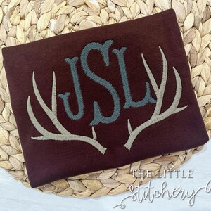 May include: A maroon fabric with a monogram of the letters "JSL" in a teal color. The monogram is centered on a pair of embroidered antlers in a beige color.