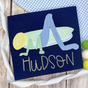 Puede incluir: Un diseño de saltamontes bordado en azul, verde y amarillo sobre una tela azul marino con el nombre "Hudson" bordado debajo.