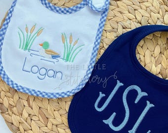 Op de afbeelding: Twee baby slabbetjes. Eén wit slabbetje met een blauwe ruitjesrand heeft een geborduurde eendenscène en de naam "Logan". Het andere slabbetje is marineblauw met de geborduurde initialen "JSL" in lichtblauw.