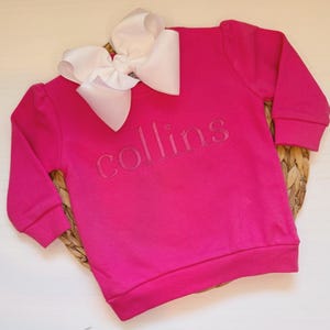 May include: A bright pink sweatshirt with the name "collins" embroidered across the chest. A large white bow is placed at the top of the sweatshirt. The sweatshirt is made of a soft material and has long sleeves.