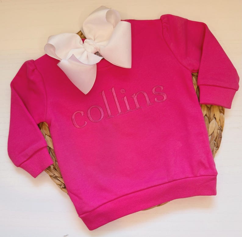May include: A bright pink sweatshirt with the name "collins" embroidered across the chest. A large white bow is placed at the top of the sweatshirt. The sweatshirt is made of a soft material and has long sleeves.