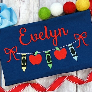 May include: A blue shirt with the name "Evelyn" embroidered on it. A red, white and green string of bunting with apples and crayons is embroidered below the name.
