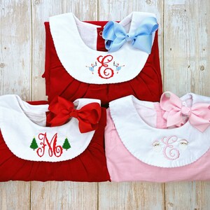 May include: Three children's dresses in red and pink, each with a white collar and a large bow. The red dresses have embroidered letters and Christmas trees, while the pink dress has an embroidered letter. The bows are in red, light blue, and pink.