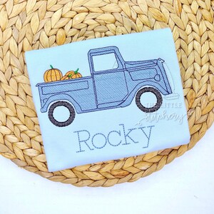 May include: Pale blue t-shirt featuring an embroidered design of a blue pickup truck with pumpkins in the back. The name "Rocky" is embroidered below. The Little Stitchory logo is in the corner.