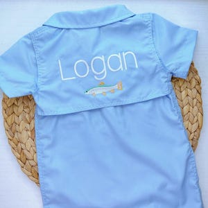 Op de afbeelding: Een lichtblauw, kort mouwig overhemd met knopen, met de naam "Logan" geborduurd op de rug, samen met een kleine geborduurde vis.