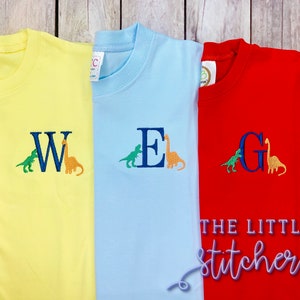 May include: Three yellow, light blue, and red t-shirts with dinosaur and letter embroidery. The shirts feature the letters W, E, and G, each with a small green dinosaur and a larger orange dinosaur beside it. The text "THE LITTLE stitchery" is on the red shirt.