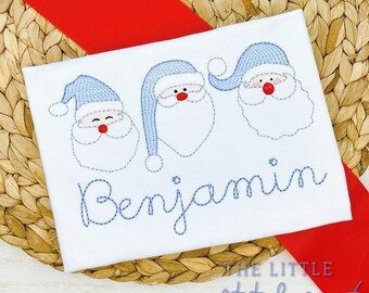 Puede incluir: Un pelele blanco con tres caras de Papá Noel bordadas en azul y el nombre "Benjamin" bordado en azul.