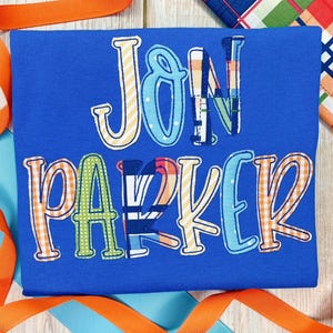 Może przedstawiać: Niebieska koszulka z imieniem "Jon Parker" wyszytym kolorowymi literami w stylu patchwork.