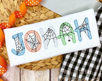 Puede incluir: Una tela blanca con el nombre "JONAH" bordado en letras de colores con telarañas y una araña en la letra "A".