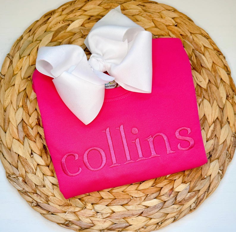 May include: A bright pink sweatshirt with the name "collins" embroidered in a matching pink thread. A large white bow is placed on top of the sweatshirt. The sweatshirt is resting on a woven, circular placemat.