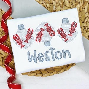 May include: White t-shirt featuring embroidered red lobsters and light blue hot air balloons. The name "Weston" is embroidered in light blue script below the design. The shirt is displayed on a woven surface.