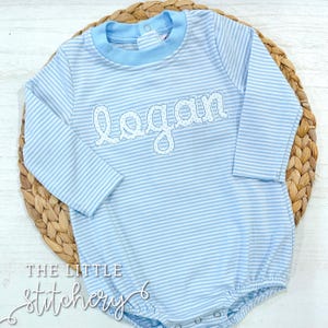 Könnte beinhalten: Hellblau-weiß gestreifter Baby-Strampler mit langen Ärmeln. Der Name "Logan" ist in weißer Schrift auf die Brust gestickt. Der Strampler hat einen hellblauen Kragen und wird auf einer gewebten Matte präsentiert. Der Text "THE LITTLE stitchery" befindet sich unten.