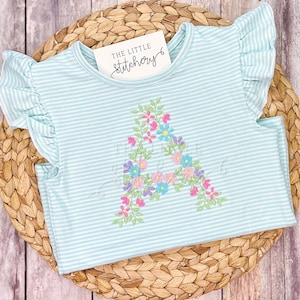 May include: A light blue and white striped t-shirt with ruffled sleeves. The shirt features an embroidered floral "A" in pink, blue, and purple. A small tag reads "THE LITTLE Stitchery".
