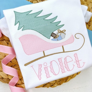 May include: A white onesie with a pink and brown embroidered design of a sleigh pulled by a Christmas tree with the name "Violet" embroidered below.
