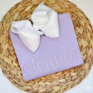 May include: A lavender sweatshirt with the name "eleanor" embroidered in a matching color. A large white bow is placed on top of the sweatshirt. The items are displayed on a woven, circular placemat.