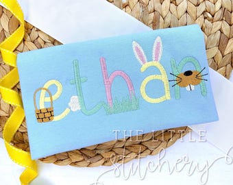 May include: A light blue t-shirt with the name "Ethan" embroidered in colourful letters. The letters are decorated with Easter-themed elements, including a bunny, a basket of eggs, and a bunny nose.