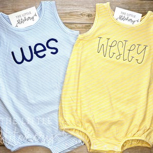 Puede incluir: Dos bodies de bebé a rayas, uno azul y blanco con el nombre "Wes" bordado, y otro amarillo y blanco con el nombre "Wesley" bordado.