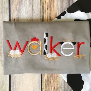 May include: A grey t-shirt with the name "Walker" embroidered in red, white and yellow. The letters are decorated with farm animal motifs, including a chicken, a cow and eggs.