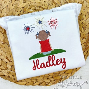 Könnte beinhalten: Weißes besticktes Shirt mit einem Feuerwerksdesign in Rot, Weiß und Blau und einem Mädchen, das die Feuerwerke beobachtet. Der Name "Hadley" ist in Rot unter dem Mädchen gestickt.