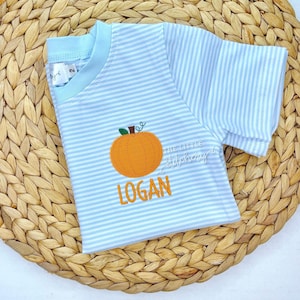 May include: A light blue and white striped t-shirt with an orange pumpkin applique and the name "LOGAN" embroidered in orange. The shirt has a light blue collar and sleeves. The text "THE LITTLE Stitchory" is also visible.