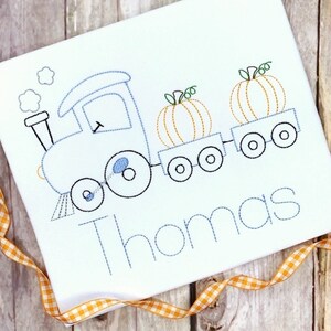 May include: A black and white outline embroidery design of a train pulling two pumpkin-shaped cars. The train has a smoke stack and wheels. The name "Thomas" is embroidered below the train.
