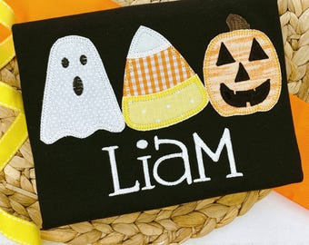 May include: A black t-shirt featuring an embroidered Halloween design with a white ghost, a candy corn with orange gingham and yellow polka dot patterns, and an orange pumpkin with a black face, with the name Liam embroidered in white.