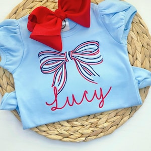 Könnte beinhalten: Ein hellblauer Baby-Strampler mit einer roten, weißen und blauen gestreiften Schleife, die auf der Vorderseite gestickt ist. Der Strampler hat auch den Namen "Lucy" in Rot unter der Schleife gestickt.