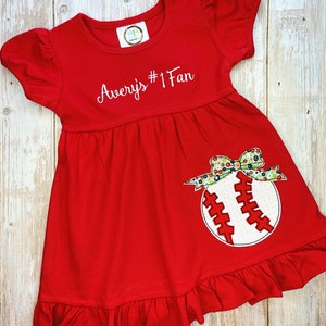 Op de afbeelding: Rode jurk zonder mouwen met een witte geborduurde baseball en de tekst "Avery's #1 Fan".
