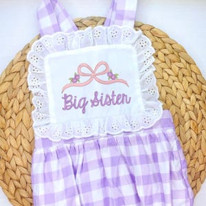 Puede incluir: Un pelele a cuadros morado claro y blanco con un babero de encaje blanco. El babero presenta el texto bordado "Big Sister" con un lazo rosa y detalles florales. El pelele tiene tirantes y un borde con volantes.