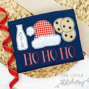 May include: A navy blue t-shirt with a red and white gingham Santa hat, a milk bottle, and two chocolate chip cookies. The text "HO HO HO" is embroidered in red and white thread.