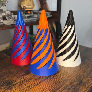 May include: Three decorative cone-shaped objects with a spiral design. One is red and blue, another is orange and blue, and the third is black and white. They are displayed on a wooden surface.