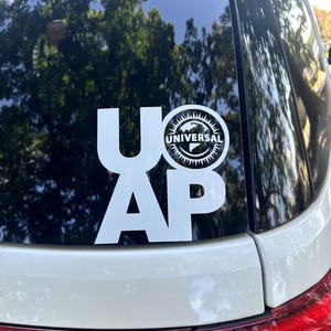 May include: White sticker on a car window featuring the letters "UAP" in a bold, sans-serif font. Above the "AP" is a circular logo with the word "UNIVERSAL" and a globe design. The sticker is affixed to a white car.