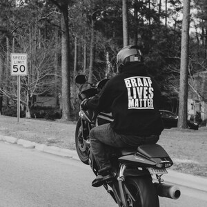 Pode incluir: Imagem a preto e branco de uma mota a fazer uma cavalinho numa estrada. O motociclista usa um capacete e um hoodie preto com o texto "BRAAP LIVES MATTER". Um sinal de limite de velocidade mostra 80 km/h.