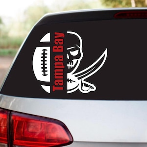 May include: A white vinyl decal featuring the Tampa Bay Buccaneers logo. The design includes a football, a skull with crossed swords, and the words "Tampa Bay" in red. The decal is applied to the rear window of a car.