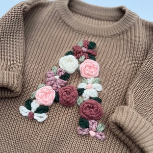 May include: A tan-colored knit sweater with a large floral "A" applique. The letter is crafted from various yarn colors, including white, pink, and burgundy, with green accents. The sweater features a ribbed knit design and long sleeves.