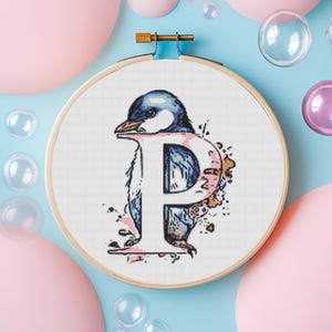 May include: A cross-stitch embroidery hoop with a white background featuring a blue and white penguin with a pink watercolor design around it. The penguin is positioned to form the letter "P".