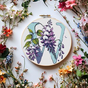 May include: A white embroidery hoop with a blue and purple floral design featuring the letter W. The hoop is surrounded by a variety of colourful flowers.
