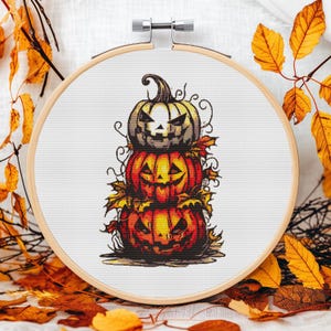 Op de afbeelding: Een kruissteekontwerp van drie op elkaar gestapelde pompoenen met uitgesneden gezichten, omringd door herfstbladeren. De borduurwerk is geplaatst in een houten hoepel, perfect voor Halloween. De pompoenen zijn oranje, geel en grijs.