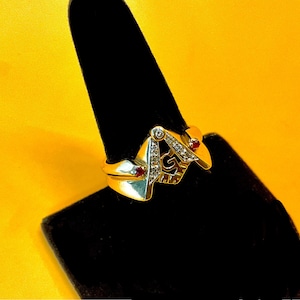 May include: A gold ring with a Masonic symbol and red and white stones.