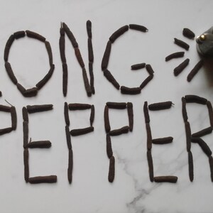 May include: The words "LONG PEPPER" are spelled out using long peppercorns on a white marble surface. A silver pepper grinder is in the top right corner of the image.