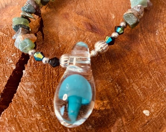 May include: A silver necklace with a clear glass pendant containing a blue glass mushroom. The necklace is made with various colored stones and black beads.