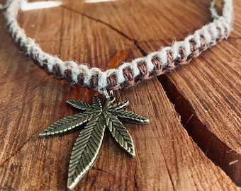 May include: A brown and white hemp necklace with a gold-toned marijuana leaf pendant.