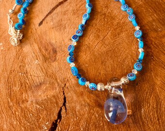 May include: A blue and white glass bead necklace with a clear glass pendant. The necklace is on a wooden surface.