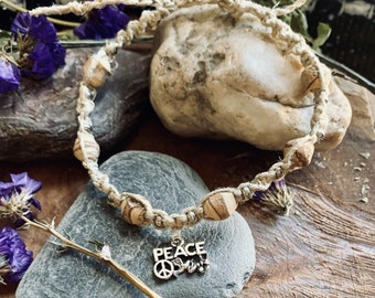 May include: A hemp bracelet with wooden beads and a peace sign charm. The bracelet is on a wooden surface with rocks and flowers.