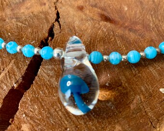 May include: A glass pendant necklace with a blue mushroom inside. The necklace is made with silver and blue beads.